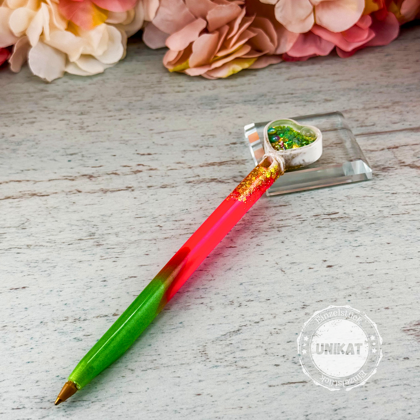 Bunter Resin-Stift mit grün-rotem Farbverlauf und Herz-Shaker-Aufsatz, querliegend auf Holzunterlage, mit sichtbarem Unikatstempel